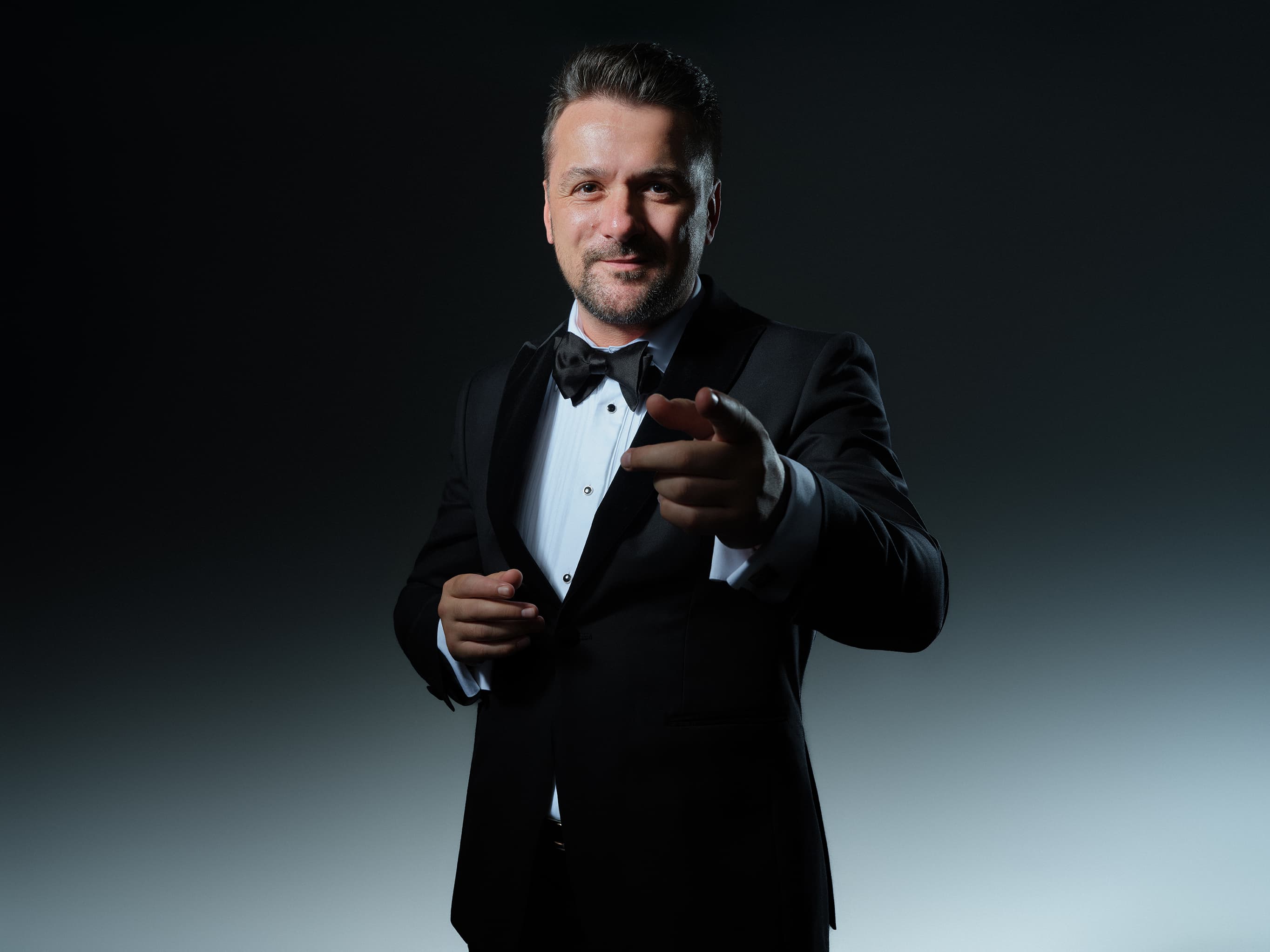 Radu Ionuț în ținută de gală, zâmbind și arătând spre cameră, pe fundal întunecat